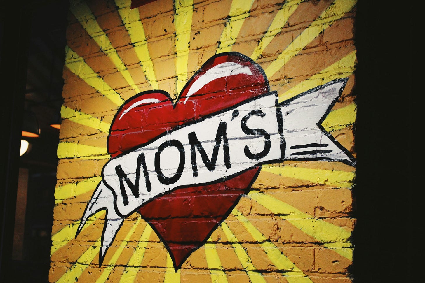 A mural of a red heart wrapped in a white banner reading MOM’S, painted on a brick wall with yellow sunburst rays